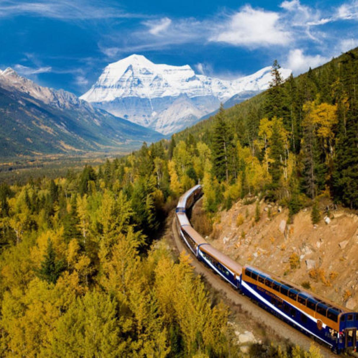 A train winds around a remote railway in the mountains, there are white capped mountains and forests in the surroundings.